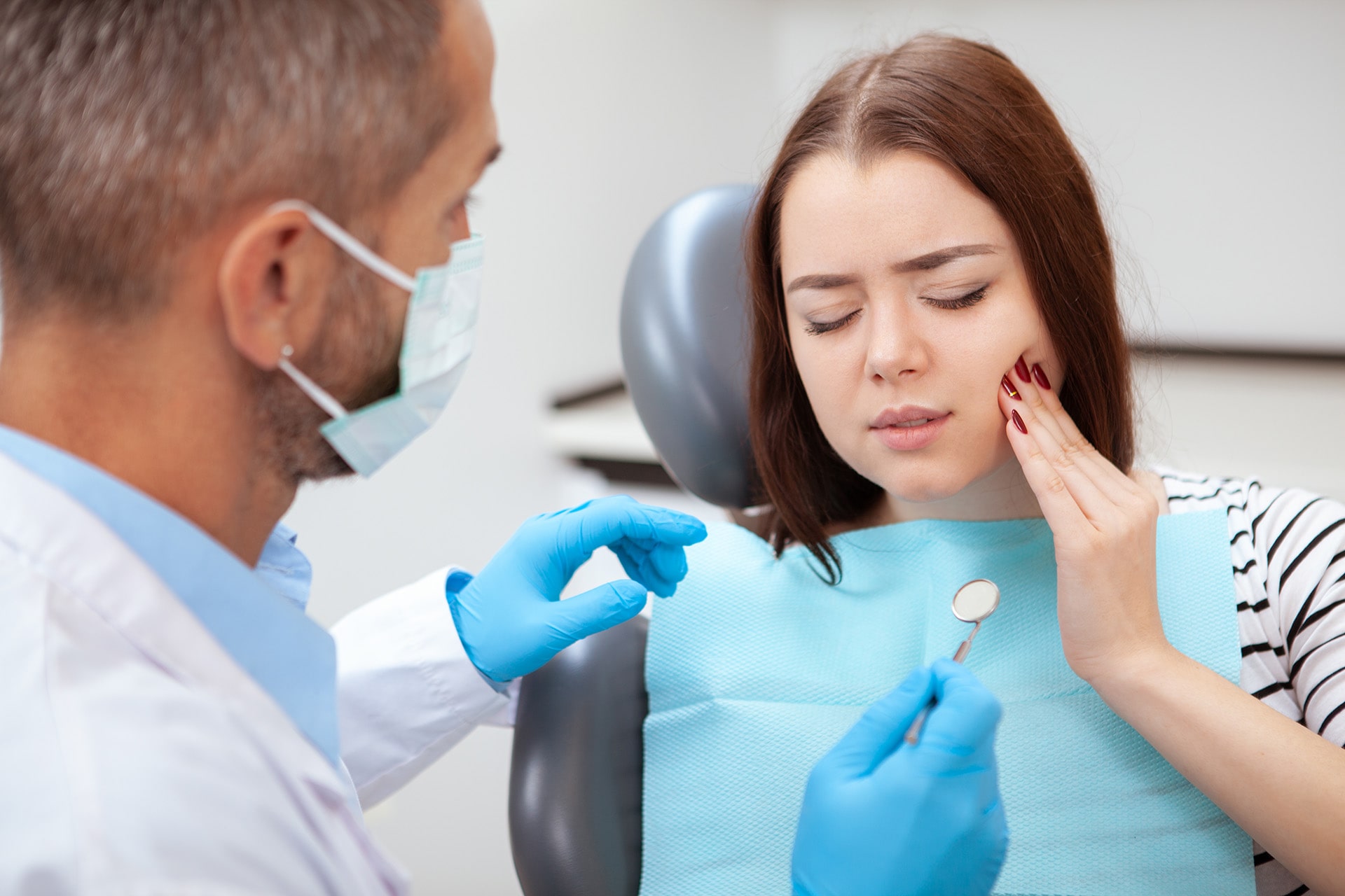 A woman experiencing severe toothache is being examined by an emergency dentist in a dental clinic. The dentist, wearing gloves, carefully checks her teeth, providing after-hours dental care to relieve the pain.